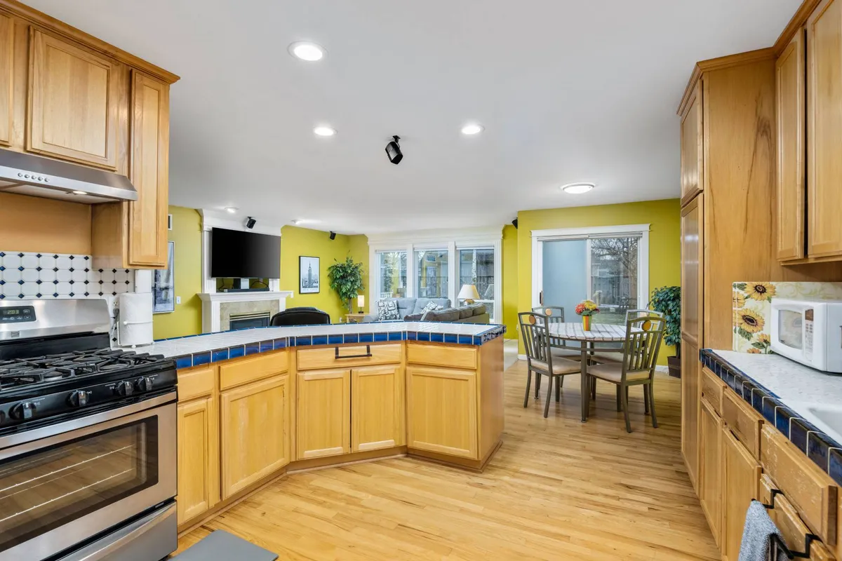 This image presents an open-concept area featuring a kitchen with a blue-tiled island, stainless steel appliances, a round dining table, and a comfortable living space— all illuminated by large windows that let in abundant Pacific Northwest sunlight. The multi-functional layout encourages cooking, dining, and relaxing in one cohesive space.