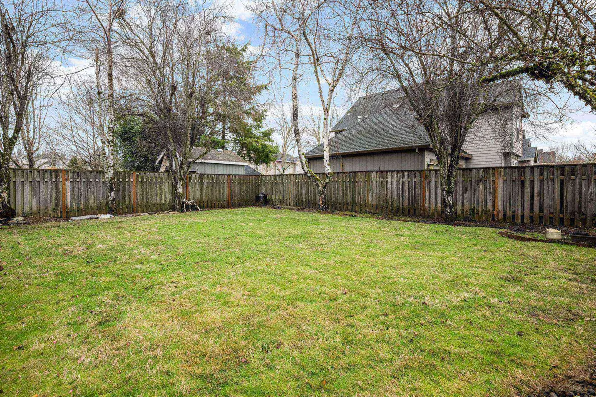 A wide shot of the fully fenced backyard, featuring a large, lush green lawn surrounded by mature trees and a visible fence perimeter. The open grassy area is perfect for children and pets to play safely, while the natural setting offers a peaceful environment for relaxation.