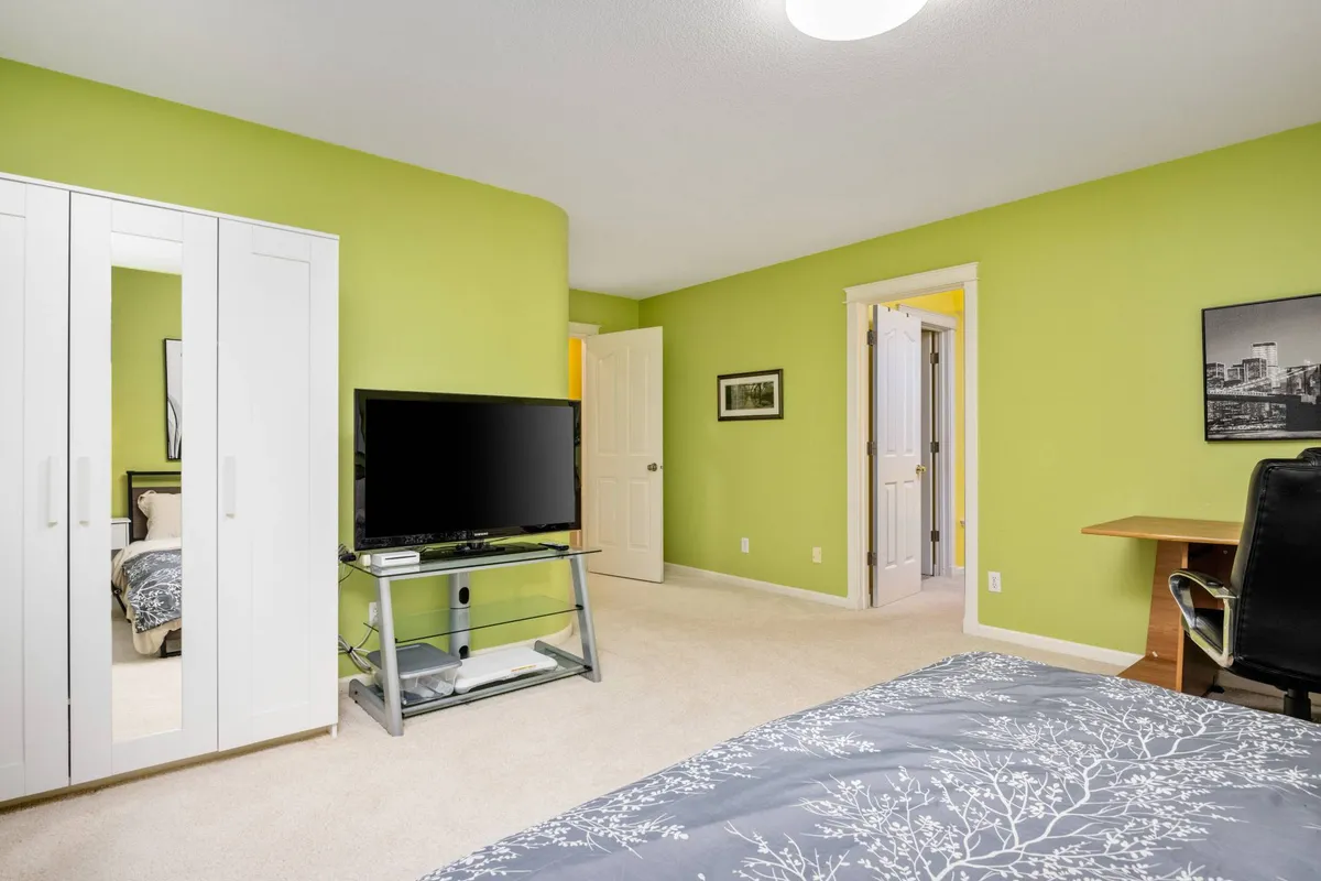 A bright, cheerful bedroom with green walls, a comfortable bed, a large TV, and a desk with a chair. The space is vibrant yet cozy, suitable for relaxing, watching TV, or working at the desk.