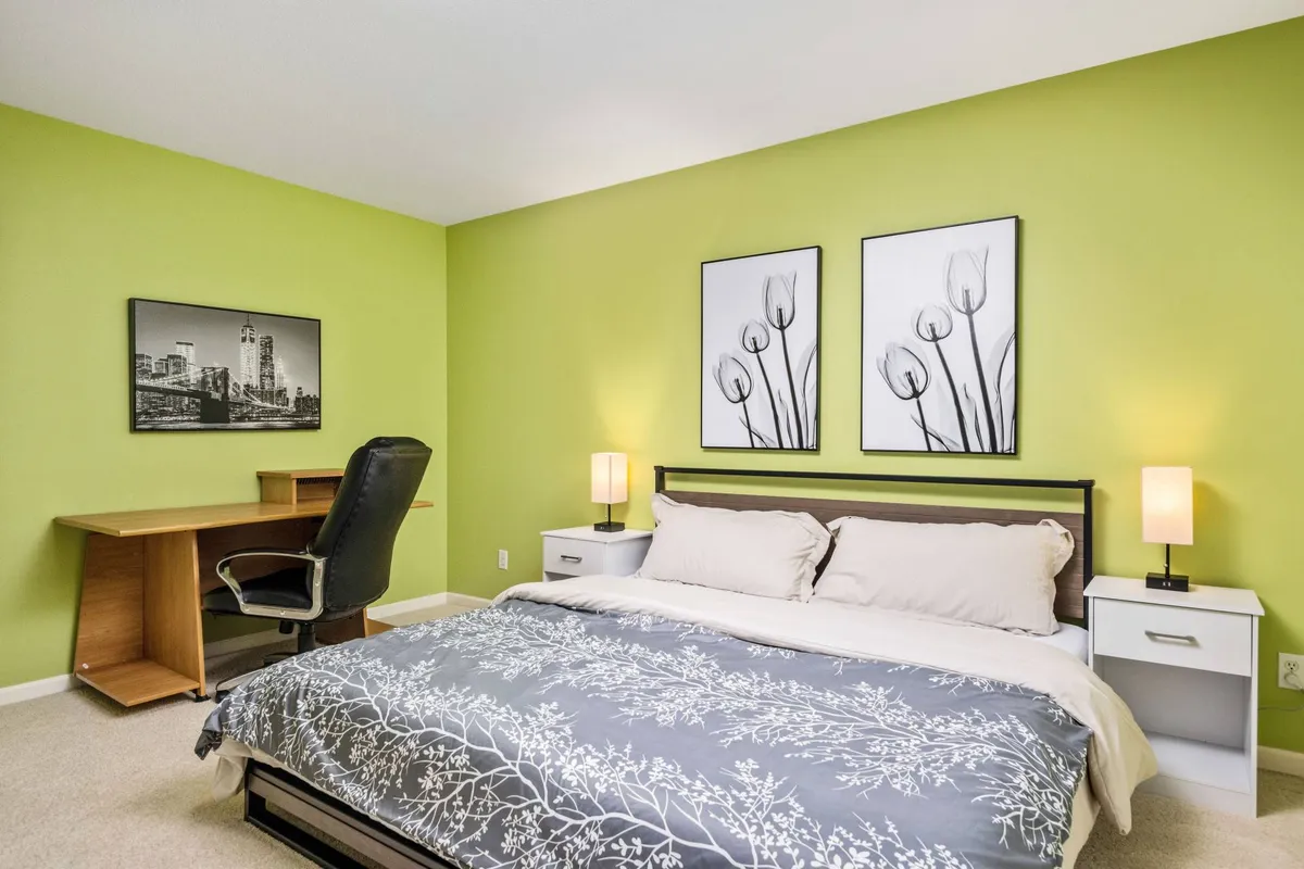 A serene bedroom featuring a bed with gray floral bedding, a calming green accent wall, a functional desk and chair, and framed artwork. The room is flooded with natural light, creating a peaceful and productive space for rest and quiet work.