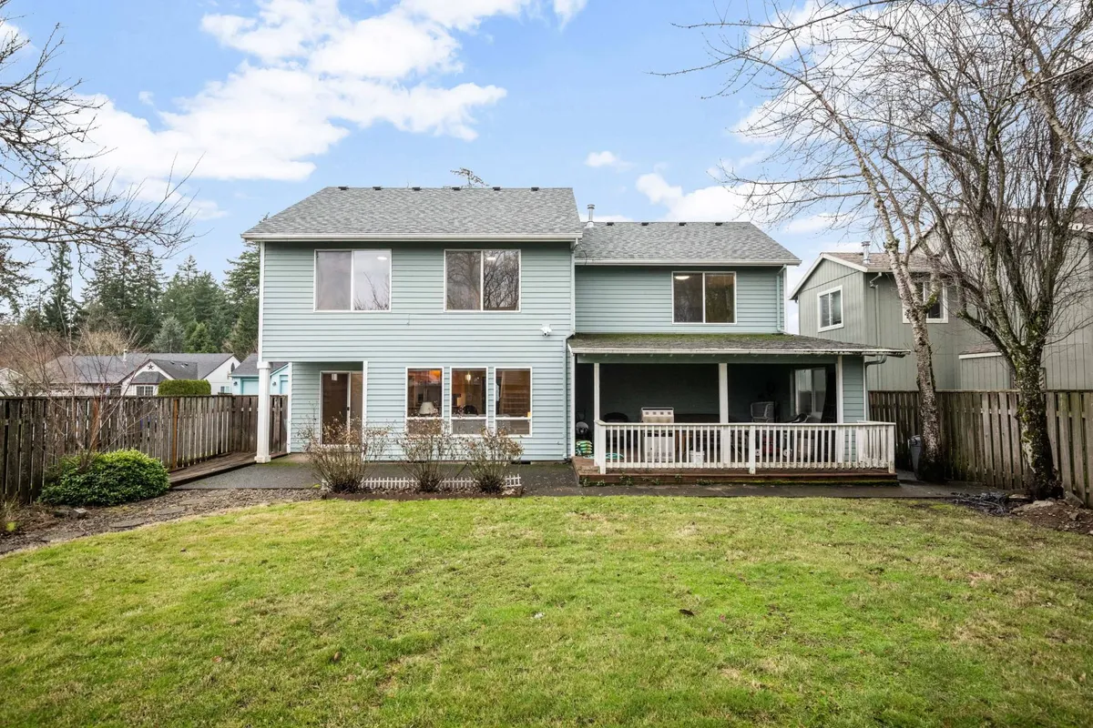 The backyard as viewed from the deck, showing the two-story house, a spacious green yard, and the deck area. The scene is set during a welcoming time of day, with hints of outdoor gatherings and the possibility of barbecues or relaxing with family. The fencing is subtly visible, reinforcing the secure, private feel.