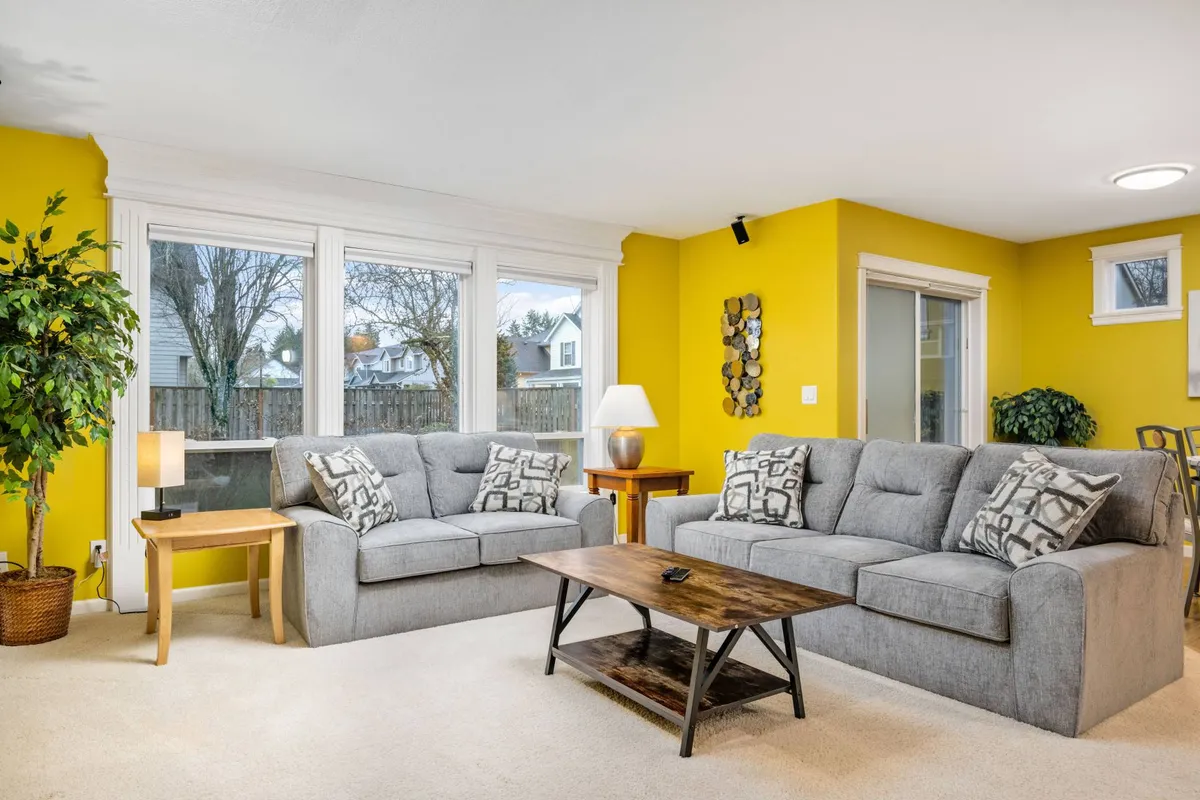 This image showcases a spacious, inviting living room with multiple sofas, a coffee table, lush plants, and large windows allowing for abundant natural light. The arrangement creates a warm atmosphere ideal for group gatherings, socializing, and family activities such as playing games or watching movies together.