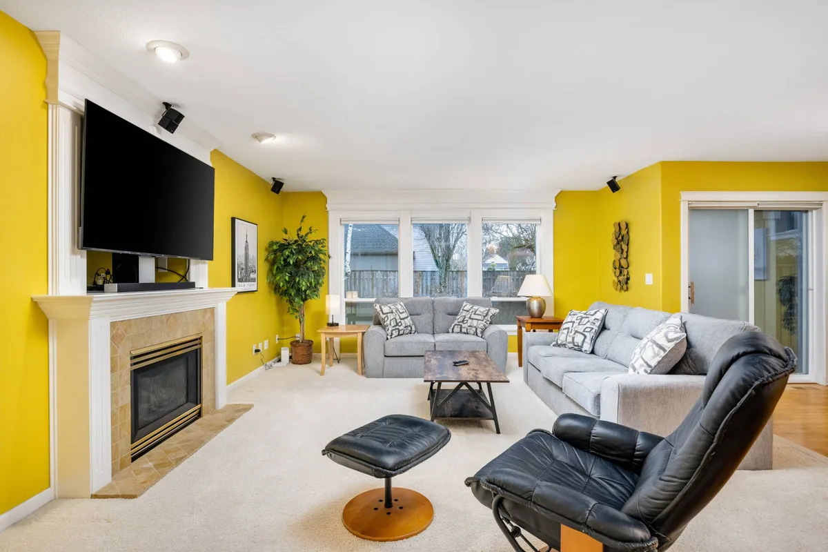 A spacious living room with yellow walls, a prominent fireplace, comfortable seating, and large windows offering views and plentiful sunlight. The setup is ideal for relaxing with family, watching TV, or sharing stories by the fire, echoing the inviting, communal atmosphere of the retreat.