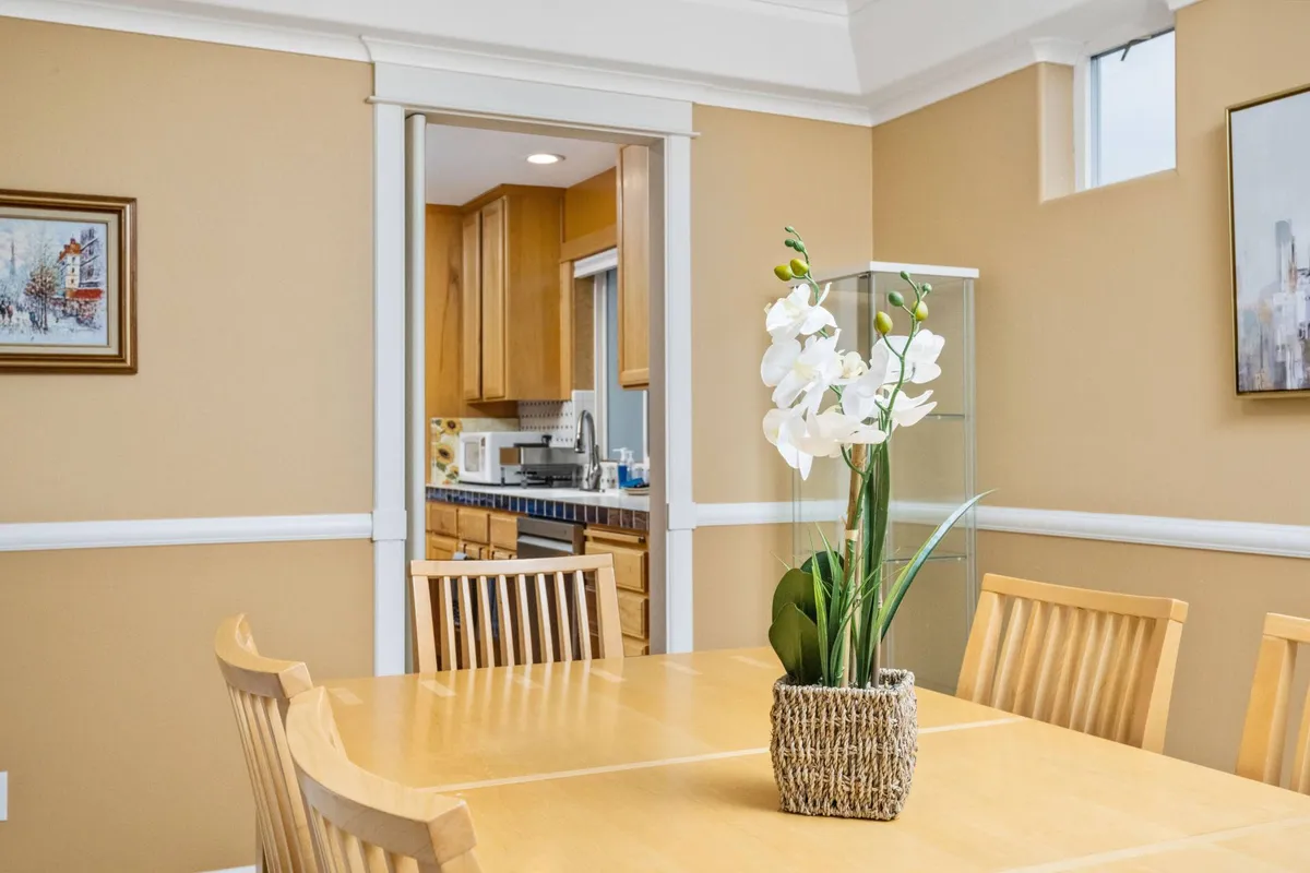 A beautifully arranged dining table featuring a vibrant flower arrangement as its centerpiece. The image includes a clear view of a doorway leading into the kitchen, framed artwork on the wall, and a sense of openness that visually connects communal spaces.